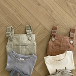 Zwei Sets Kindergarderobe liegen auf einem Holzboden. Das linke Set besteht aus einem beigen Bebe Holic Pigment Latzhose Body mit verstellbaren Trägern, einem grauen Shirt und passenden Socken. Die rechte Kindergarderobe besteht aus einem braunen Overall, einem cremefarbenen Shirt mit Smiley und hellgrauen Socken.