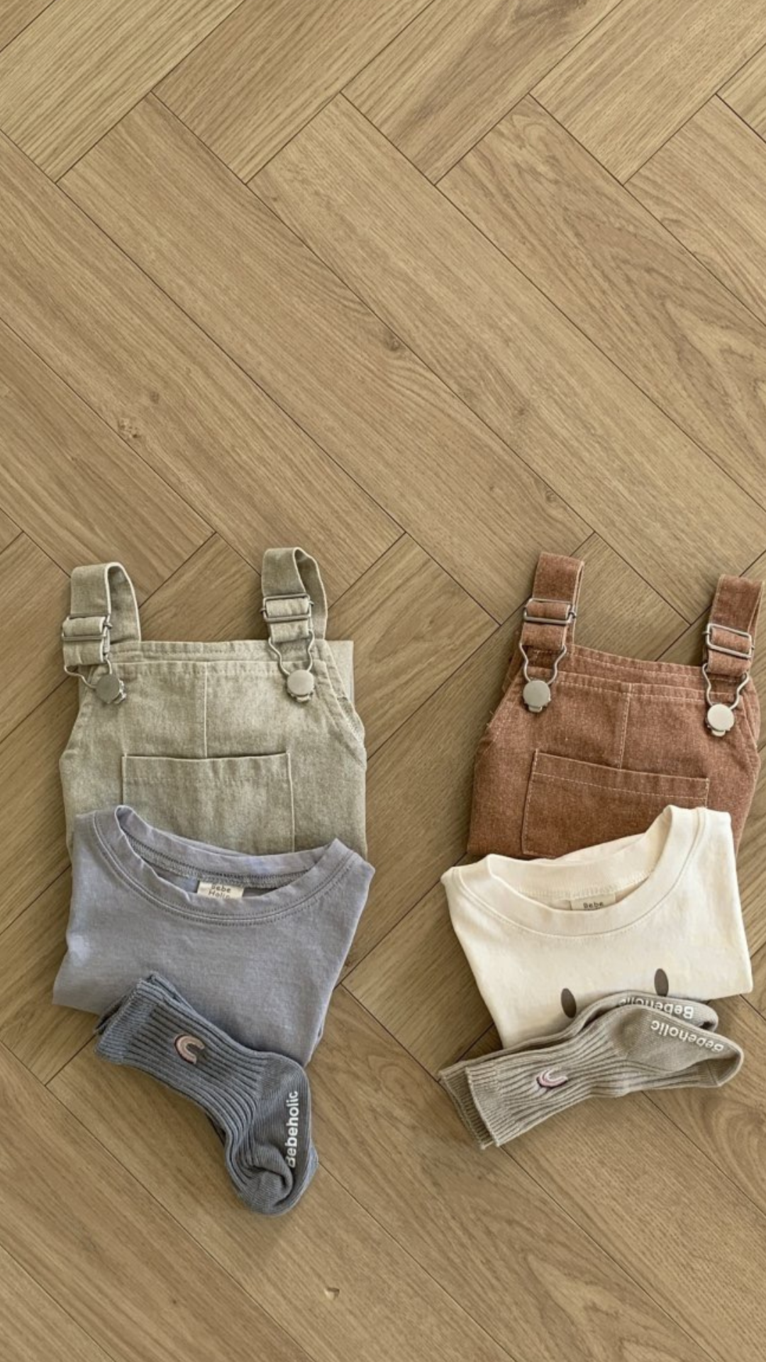 Zwei Sets Kindergarderobe liegen auf einem Holzboden. Das linke Set besteht aus einem beigen Bebe Holic Pigment Latzhose Body mit verstellbaren Trägern, einem grauen Shirt und passenden Socken. Die rechte Kindergarderobe besteht aus einem braunen Overall, einem cremefarbenen Shirt mit Smiley und hellgrauen Socken.