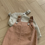 Zwei Paar Kinderoveralls liegen auf einem Holzboden. Das Paar im Vordergrund, gefertigt von Bebe Holic, ist ein Pigment Dungarees Body Suit in einem sanften Braunton mit verstellbaren Trägern und einer Vordertasche. Das Paar im Hintergrund ist hellbeige mit ebenso verstellbaren Trägern. Sonnenlicht fällt von der oberen linken Ecke ein.