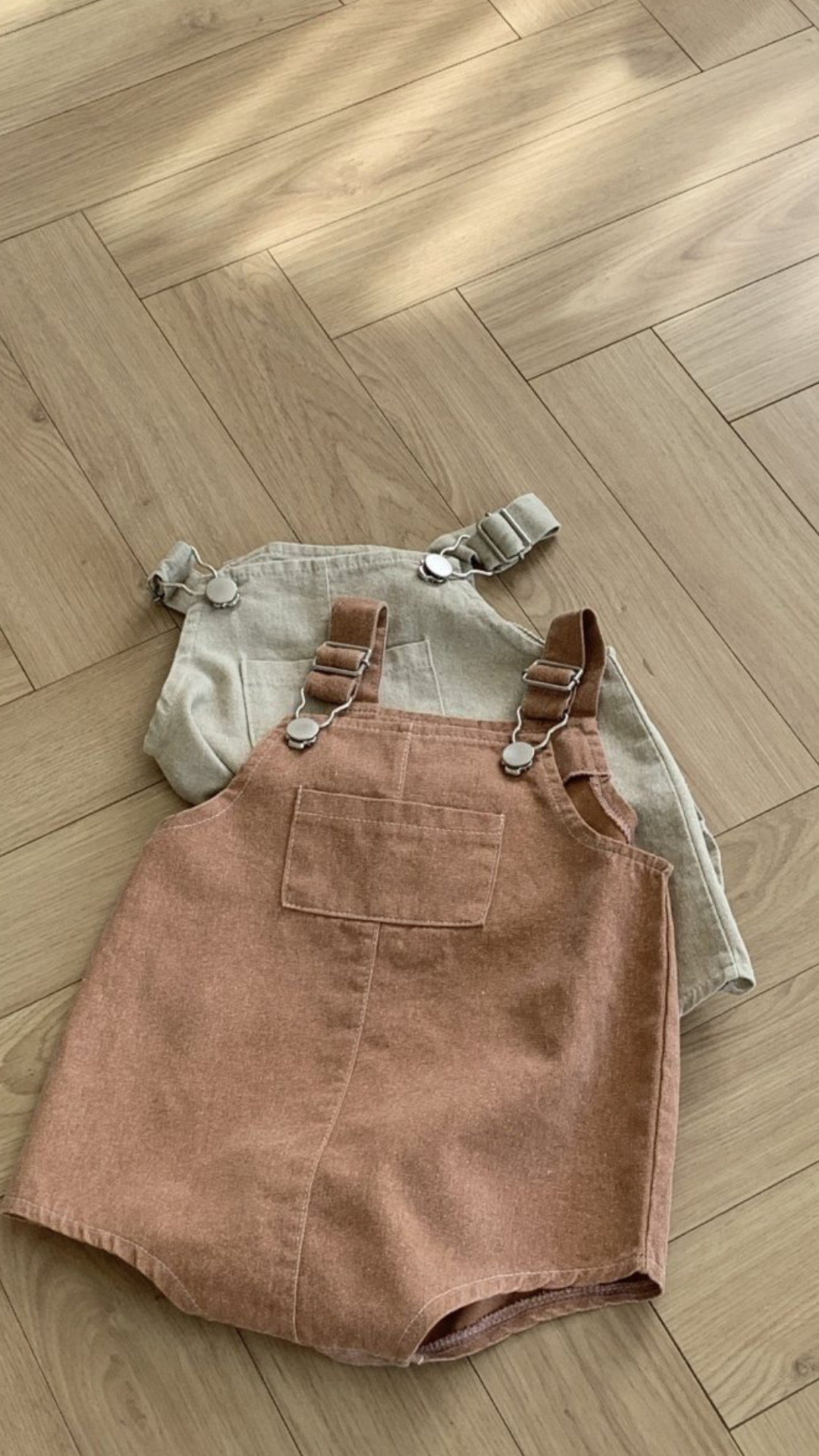 Zwei Paar Kinderoveralls liegen auf einem Holzboden. Das Paar im Vordergrund, gefertigt von Bebe Holic, ist ein Pigment Dungarees Body Suit in einem sanften Braunton mit verstellbaren Trägern und einer Vordertasche. Das Paar im Hintergrund ist hellbeige mit ebenso verstellbaren Trägern. Sonnenlicht fällt von der oberen linken Ecke ein.