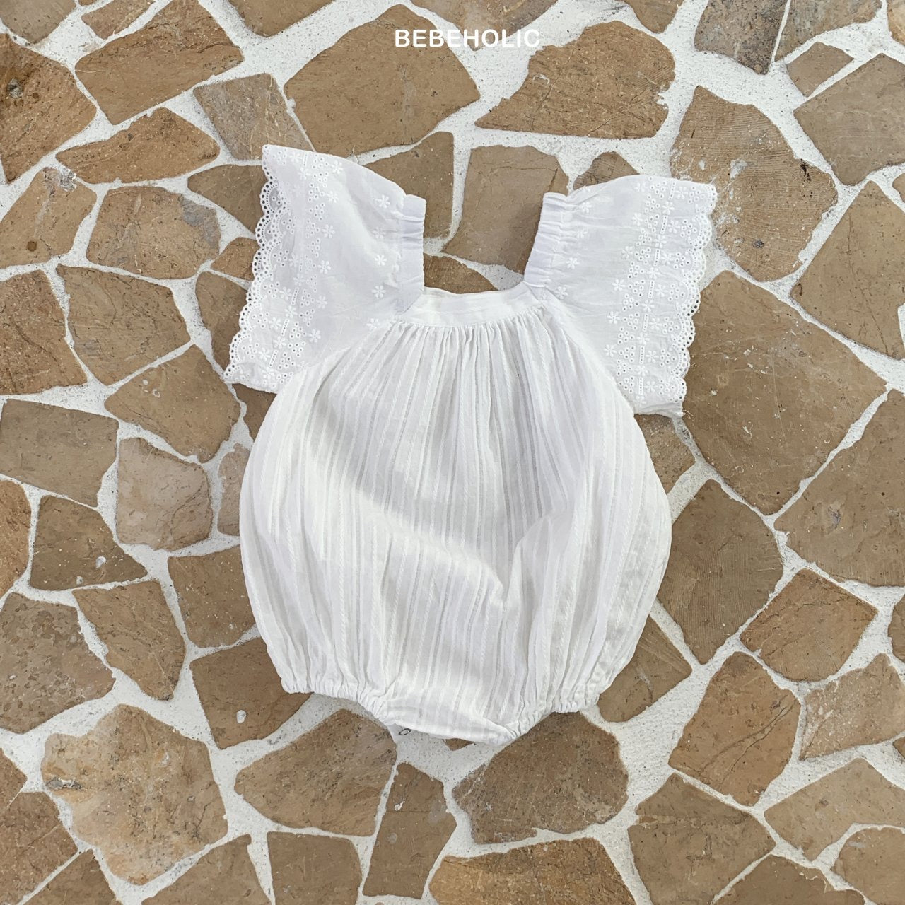 Ein weißer Wing Body Suit von Bebe Holic mit Spitzenschulterdetails liegt flach auf einem Mosaiksteinpflaster. Der Stoff wirkt leicht und eignet sich für ein Sommeroutfit.