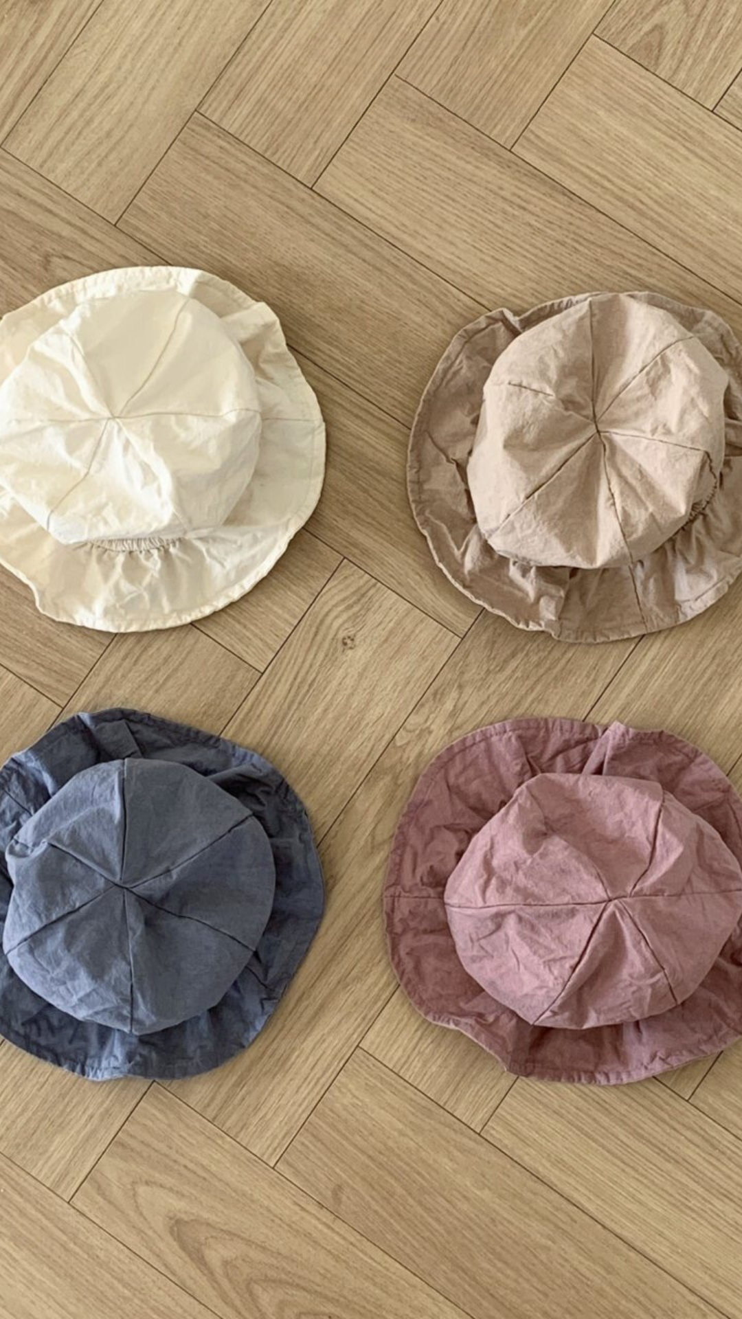 Vier zerknitterte Crunch Bucket Hats liegen in einem 2x2-Raster auf einem Holzboden. Die Hüte in einer natürlichen Farbpalette aus Beige, Hellbraun, Blau und Rosa haben jeweils eine zerknitterte Textur, die ihnen ein lässiges Aussehen verleiht. Perfekt für die Kleinen von Bebe Holic.