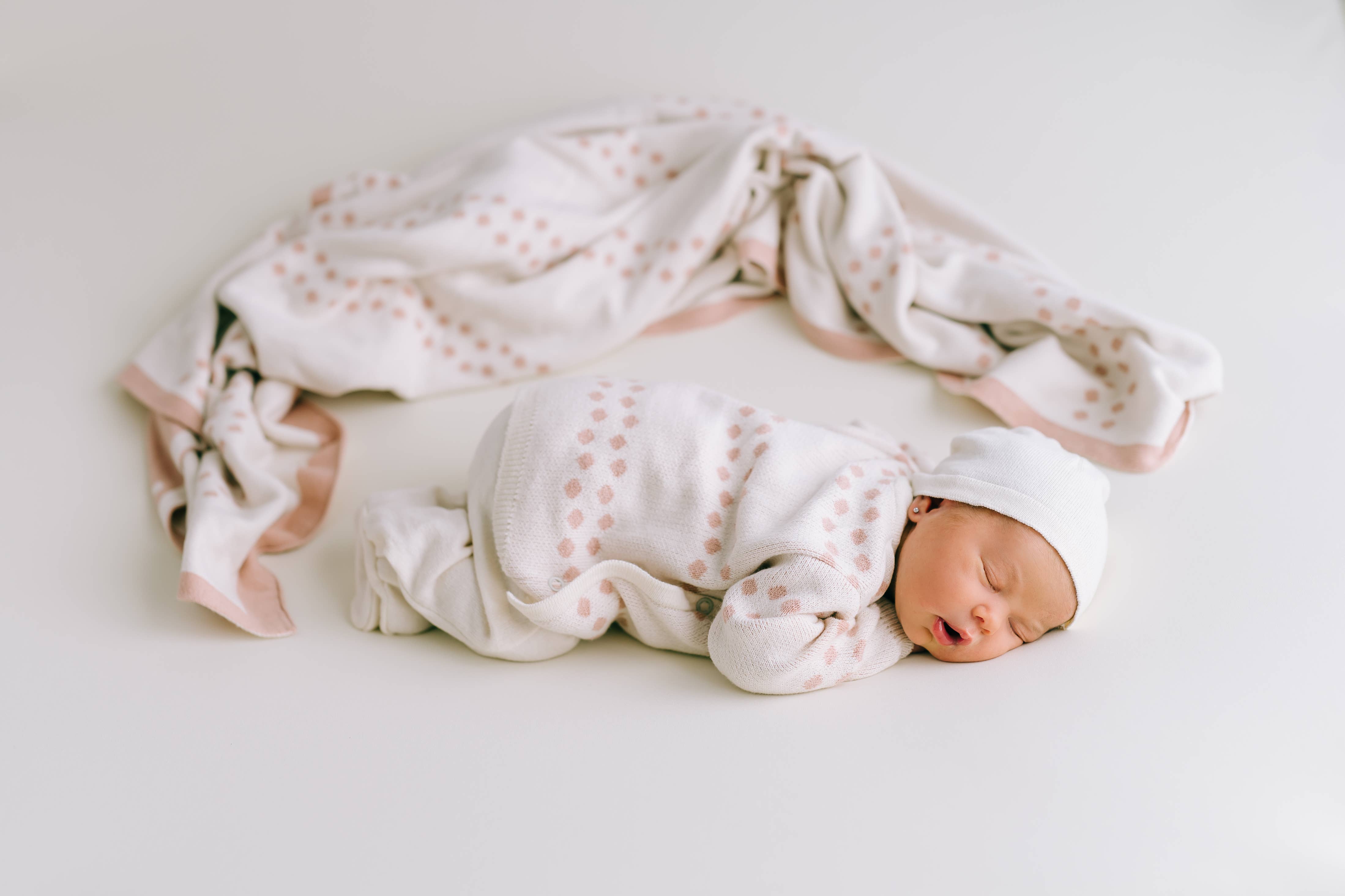 Ein Neugeborenes schläft friedlich auf einer weißen Oberfläche. Das Baby trägt ein weißes Outfit mit hellbraunem Muster und eine passende Mütze aus antiallergischer Baumwolle. Eine weiche, gemusterte Decke liegt hinter dem Baby und verleiht der Szene eine gemütliche Note. Das entzückende Ensemble ist Teil des "Newborn Pack Punkte rosa: 0 -3 Monat (56 cm)" von micu micu.