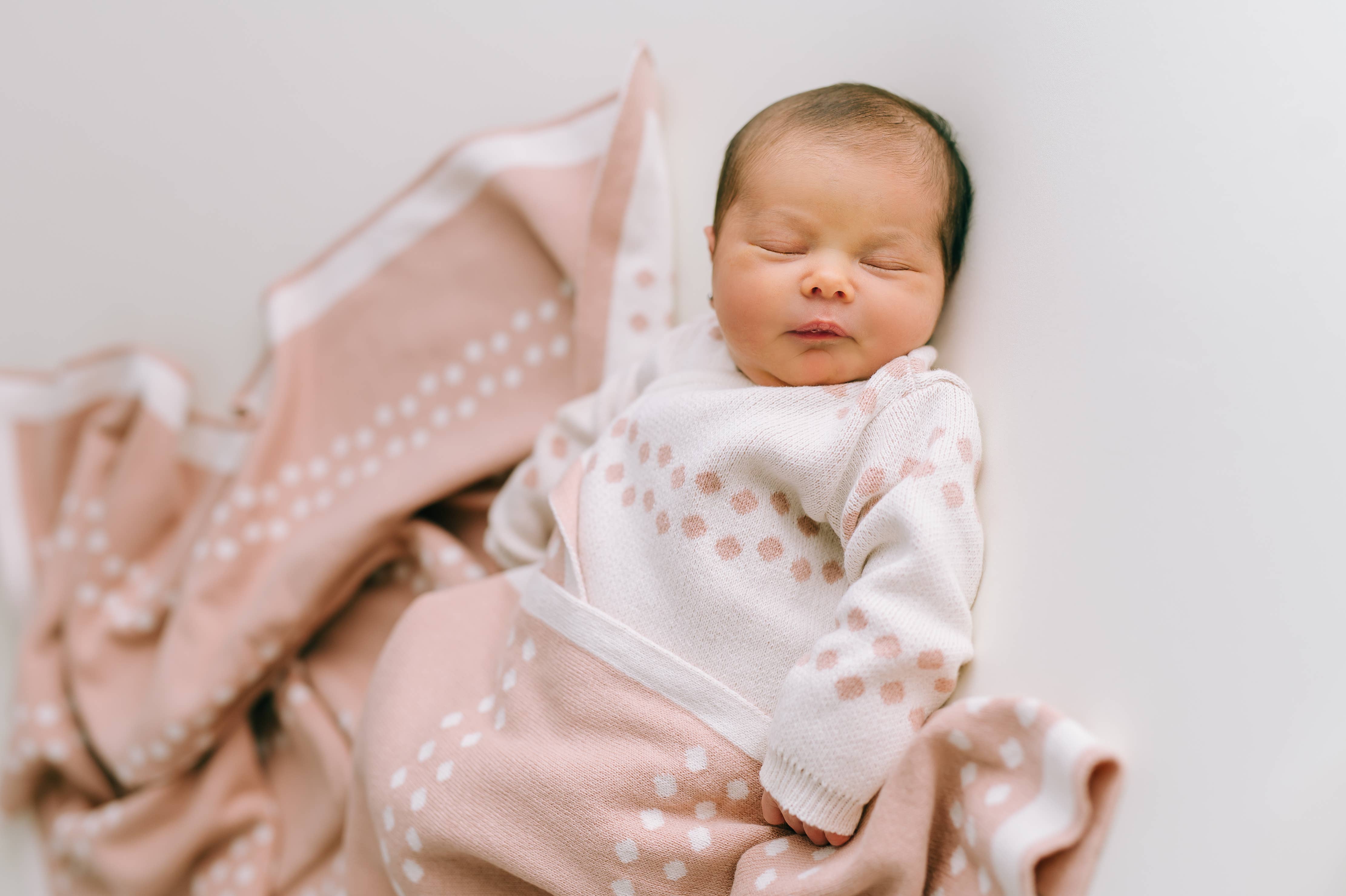 Ein Neugeborenes schläft friedlich, eingehüllt in eine weiche rosa-weiße Decke aus antiallergischer Baumwolle, die zum Babyoutfit passt. Das Baby liegt auf einem weißen Hintergrund und die Szene strahlt Wärme und Ruhe aus. Die Decke ist Teil des Newborn Pack Punkte rosa: 0 -3 Monat (56 cm) von micu micu.