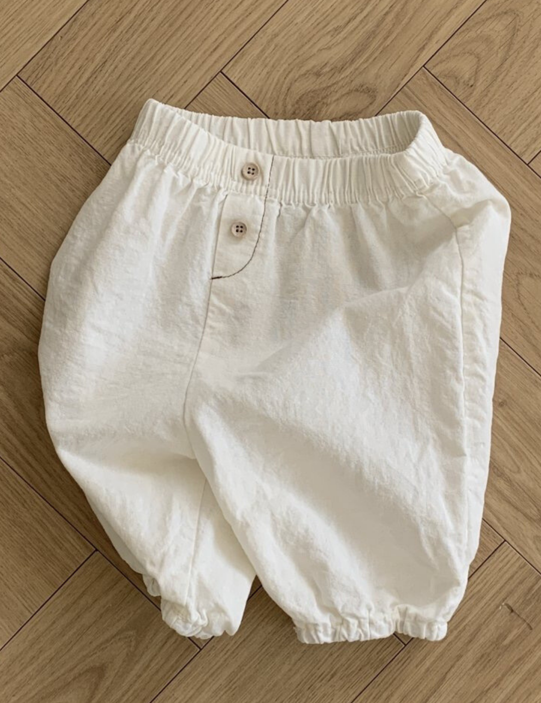 Auf einem Holzboden ist eine weiße Bebe Holic Natural Pants mit elastischem Bund und Knopfverschluss abgebildet. Die Hose hat eine lockere, bequeme Passform mit elastischen Beinöffnungen und ist damit ein vielseitiges Must-have für die Garderobe jedes Babys.