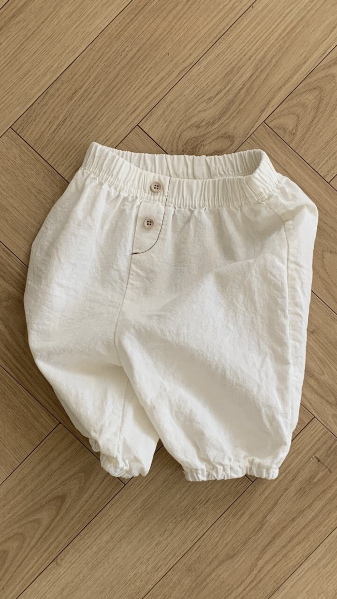 Auf einem Holzboden ist eine weiße Bebe Holic Natural Pants mit elastischem Bund und Knopfverschluss abgebildet. Die Hose hat eine lockere, bequeme Passform mit elastischen Beinöffnungen und ist damit ein vielseitiges Must-have für die Garderobe jedes Babys.