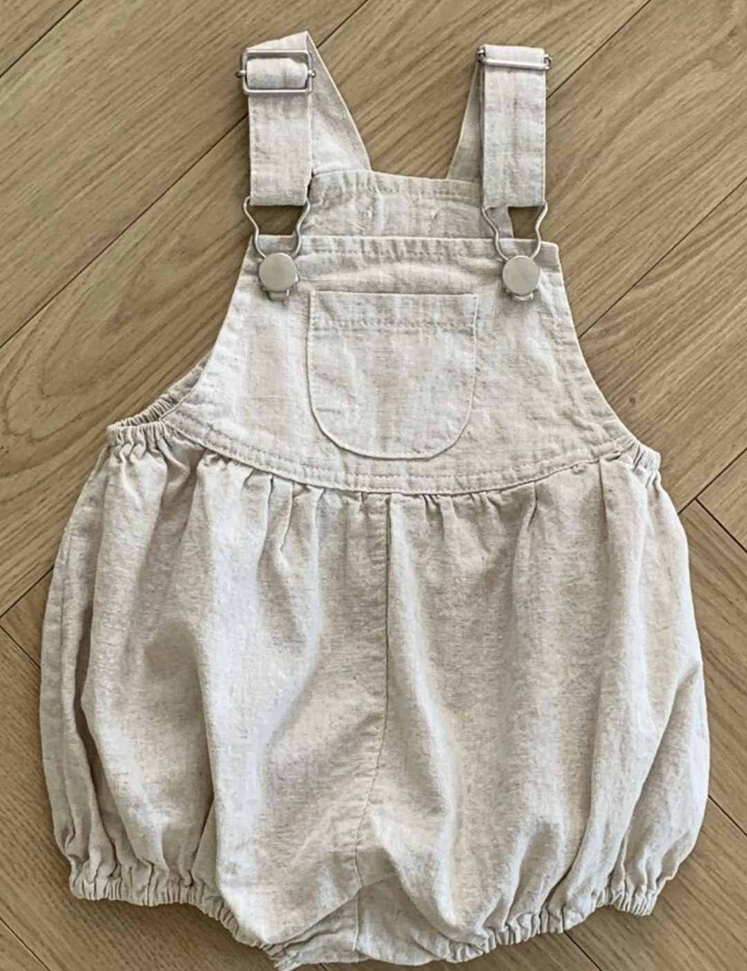 Ein beiger Baby-Strampler aus atmungsaktiver Baumwolle mit verstellbaren Trägern und Metallknopfverschlüssen auf Holzboden. Diese Bebe Holic Leinen-Latzhose mit Schnalle hat eine Vordertasche, eine geraffte Taille und locker sitzende Shorts für mehr Komfort.