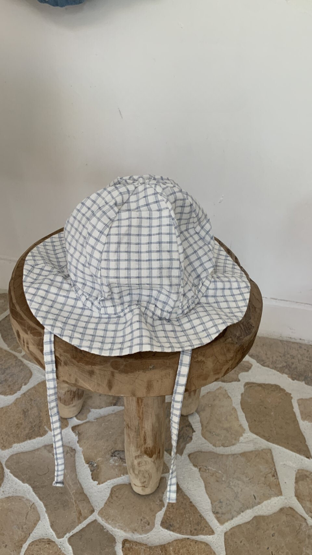 Ein weiß-blau karierter Yogurt ST Bucket Hat von Bebe Holic mit Kinnriemen sitzt auf einem kleinen, rustikalen Holzhocker. Hergestellt aus atmungsaktiven Materialien bietet er idealen Sonnenschutz. Der Hintergrund besteht aus einer weißen Wand und einem Boden mit einem beige-weißen Steinfliesenmuster.