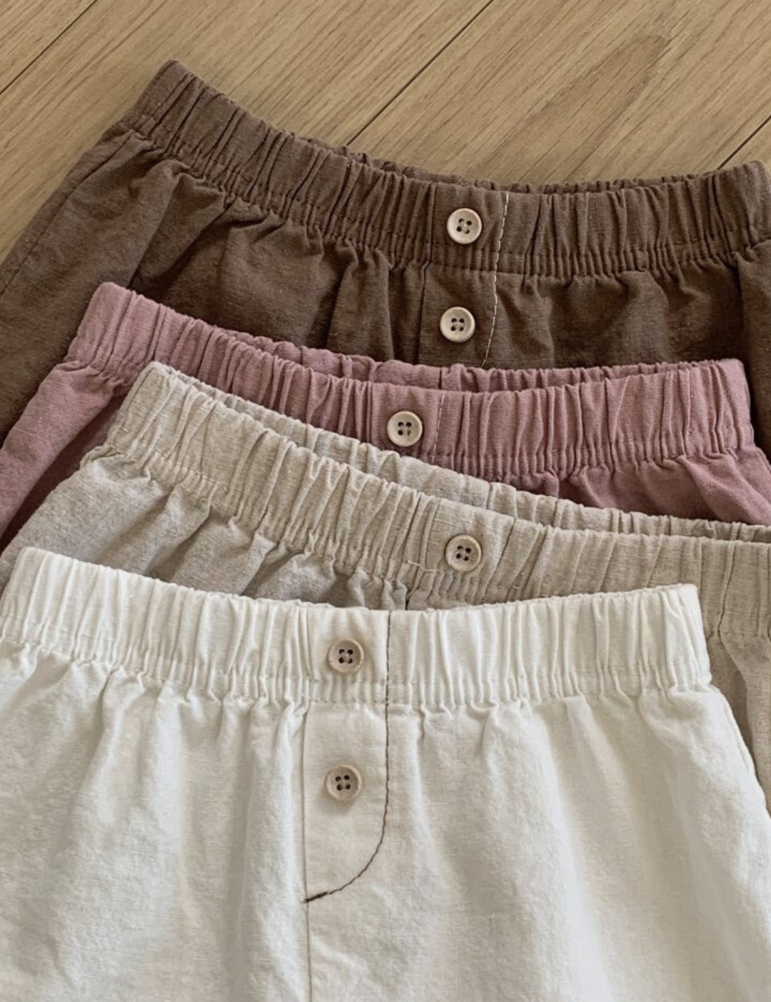 Vier Paar Bebe Holic Natural Pants mit elastischer Taille liegen auf einer Holzfläche. Die Shorts sind in verschiedenen Farben erhältlich: Braun, Mauve, Beige und Creme, von oben bis unten. Jedes Paar hat vorne in der Mitte einen Knopfverschluss – ein vielseitiges Must-have für jede Garderobe.