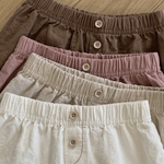 Vier Paar Bebe Holic Natural Pants mit elastischer Taille liegen auf einer Holzfläche. Die Shorts sind in verschiedenen Farben erhältlich: Braun, Mauve, Beige und Creme, von oben bis unten. Jedes Paar hat vorne in der Mitte einen Knopfverschluss – ein vielseitiges Must-have für jede Garderobe.