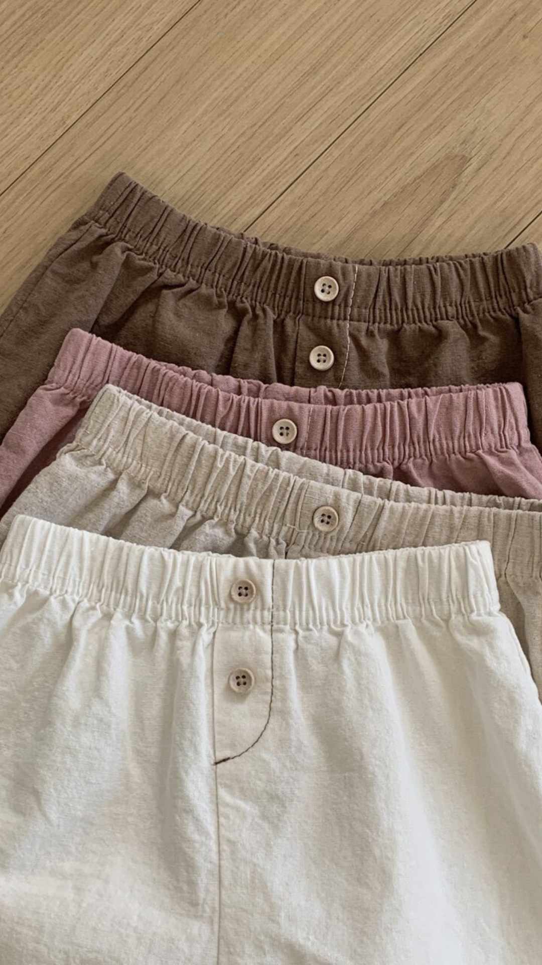 Vier Paar Bebe Holic Natural Pants mit elastischer Taille liegen auf einer Holzfläche. Die Shorts sind in verschiedenen Farben erhältlich: Braun, Mauve, Beige und Creme, von oben bis unten. Jedes Paar hat vorne in der Mitte einen Knopfverschluss – ein vielseitiges Must-have für jede Garderobe.