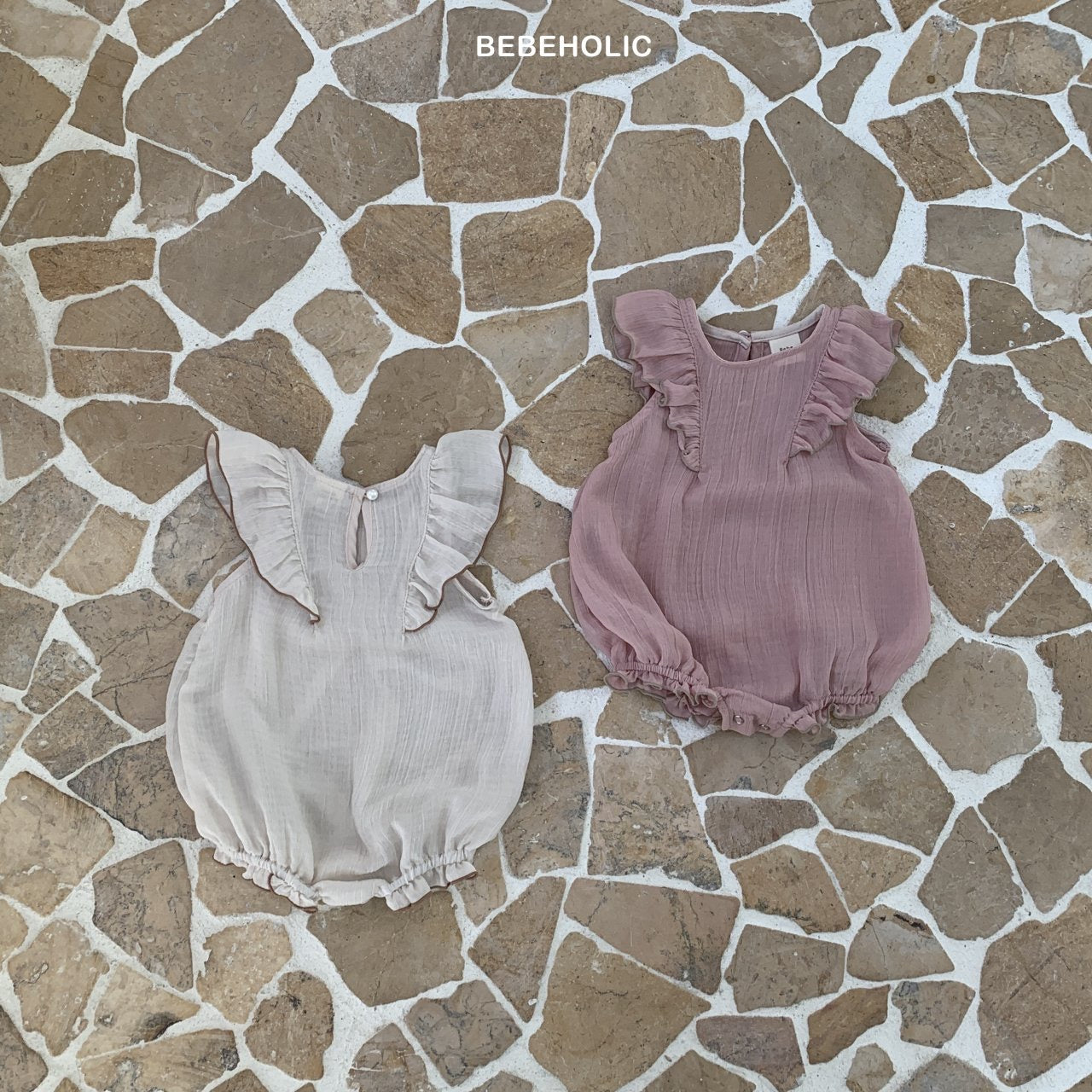 Zwei Lily Wing Body Suits auf einem Mosaikfliesenboden. Einer ist grau und der andere staubrosa, beide mit gerüschten Ärmeln und gerafftem Ausschnitt. Der Text „Bebe Holic Strampler