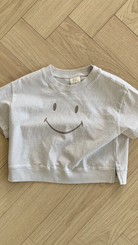 Ein weißes, kurzes T-Shirt namens Smile Tee aus der Bebe Holic-Kollektion mit einem großen Smiley auf der Vorderseite liegt auf einem hellen Holzboden mit Fischgrätenmuster. Das Smile Tee besteht aus weicher Baumwolle, hat kurze Ärmel und ein lässiges Design.