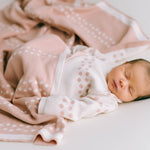 Ein Neugeborenes schläft friedlich auf einer hellen Unterlage, eingehüllt in eine rosa Decke mit weißen Mustern aus antiallergischer Baumwolle. Das Baby trägt ein weißes Outfit mit rosa Punkten, das zur Decke passt und eine weiche und heitere Atmosphäre schafft. Das verwendete Produkt ist das Newborn Pack Punkte rosa: 0 -3 Monat (56 cm) von micu micu.