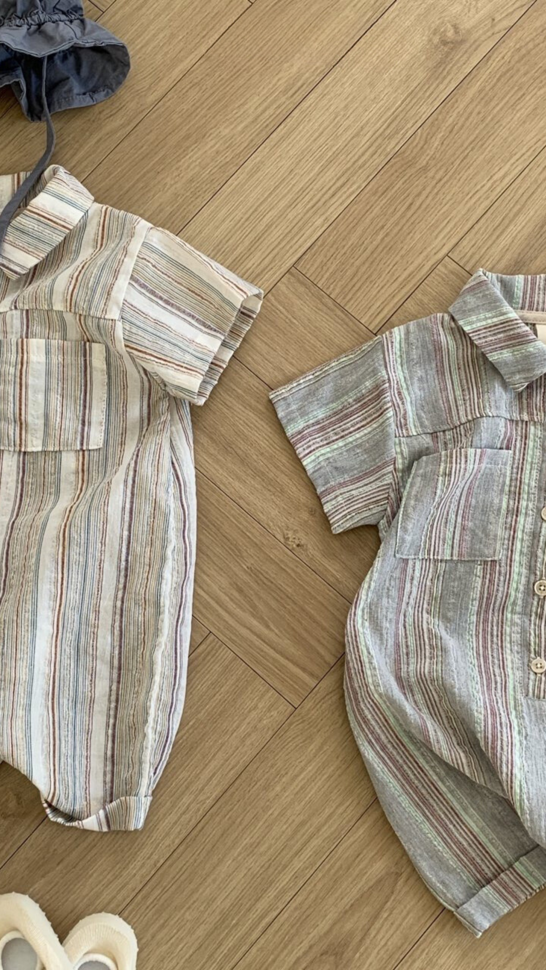 Zwei gestreifte Aurora-Bodys von Bebe Holic, einer überwiegend beige und einer überwiegend grau, liegen auf einem hellen Holzboden. Daneben liegen Babyschuhe und passende Hüte in Marine mit Schnüren. Die Outfits haben kurze Ärmel und Vordertaschen.