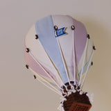 Ein kleiner dekorativer Ballon – Beige, Mint, Grau und Lila | Einzigartiges Heißluftballon-Ornament für Ihr Zuhause mit pastellblauen, weißen und rosa Paneelen. Der Korb besteht aus braunem Weidengeflecht und ist mit weißen Schnüren mit dem Ballon verbunden. Auf dem Ballon steht „SUPER 500 BALLOON“. Diese vielseitige Dekoration von Superballoon fällt vor einer schlichten, hellen Wand auf.