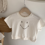 Ein minimalistisches weißes kurzes T-Shirt mit dem Namen Bebe Holic Smile Tee und einem einfachen Smiley-Design hängt auf einem hölzernen Kleiderbügel vor einer weißen Wand. Das Shirt aus weicher Baumwolle ist kurzärmlig. Daneben liegen karierter Stoff und eine Holzstufe, die eine ruhige und organisierte Atmosphäre schaffen.