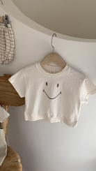 Ein minimalistisches weißes kurzes T-Shirt mit dem Namen Bebe Holic Smile Tee und einem einfachen Smiley-Design hängt auf einem hölzernen Kleiderbügel vor einer weißen Wand. Das Shirt aus weicher Baumwolle ist kurzärmlig. Daneben liegen karierter Stoff und eine Holzstufe, die eine ruhige und organisierte Atmosphäre schaffen.