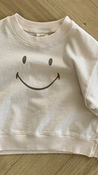 Ein kleines, hellrosa Bebe Holic Smile Tee liegt auf einem Holzboden. Das Sweatshirt hat auf der Vorderseite ein großes, einfaches schwarzes Smiley-Design und ist aus weicher Baumwolle.