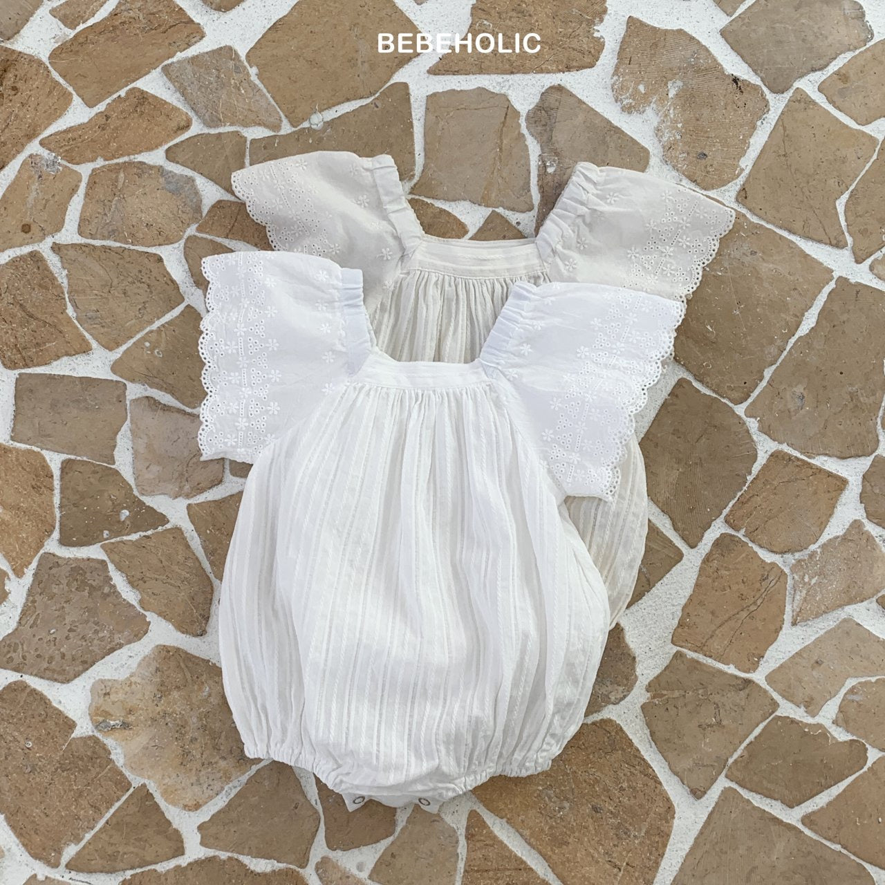 Weißes Babykleid von Bebe Holic mit Spitzendetails, flach auf einer geometrisch gemusterten Fliesenoberfläche. Das Kleidungsstück hat Puffärmel und ist für Kleinkinder konzipiert.