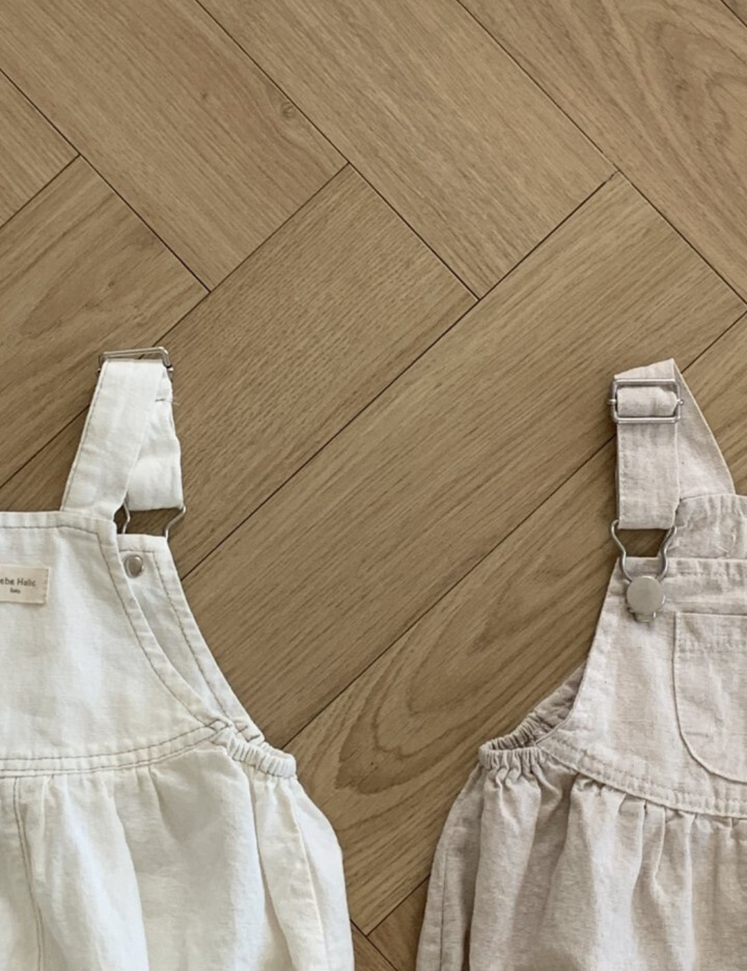 Zwei Bebe Holic Leinen-Latzhosen mit Schnallen liegen auf einem hellen Fischgrätmusterboden aus Holz. Der linke Overall aus atmungsaktiver Baumwolle ist weiß mit einer dekorativen Kerbe am Latz, und der rechte ist beige mit Falten an der Taille. Beide verfügen über Metallschnallen und verstellbare Träger.