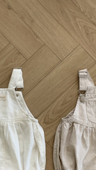 Zwei Bebe Holic Leinen-Latzhosen mit Schnallen liegen auf einem hellen Fischgrätmusterboden aus Holz. Der linke Overall aus atmungsaktiver Baumwolle ist weiß mit einer dekorativen Kerbe am Latz, und der rechte ist beige mit Falten an der Taille. Beide verfügen über Metallschnallen und verstellbare Träger.