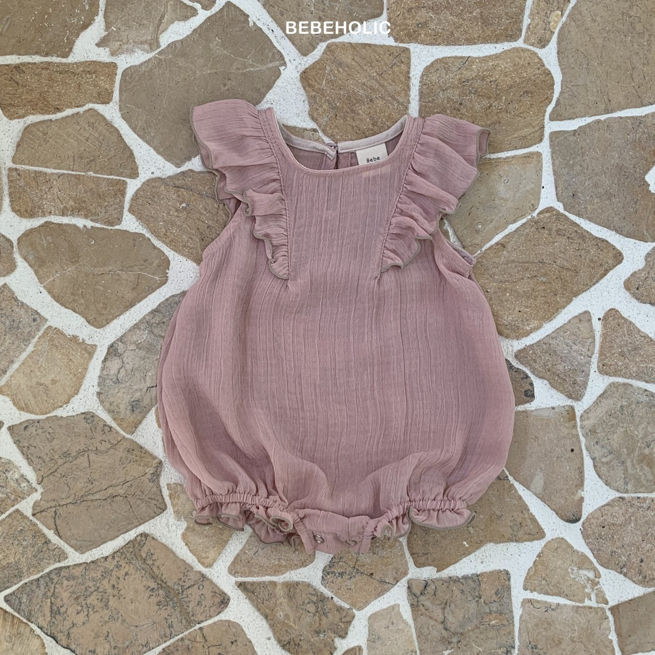 Ein rosa Lily Wing Bodysuit mit romantischen Rüschen und elastischen Beinausschnitten auf einer Pflastersteinoberfläche. Der Strampler ist aus strukturiertem Stoff und hat ein kleines Etikett mit der Marke „Bebe Holic“.