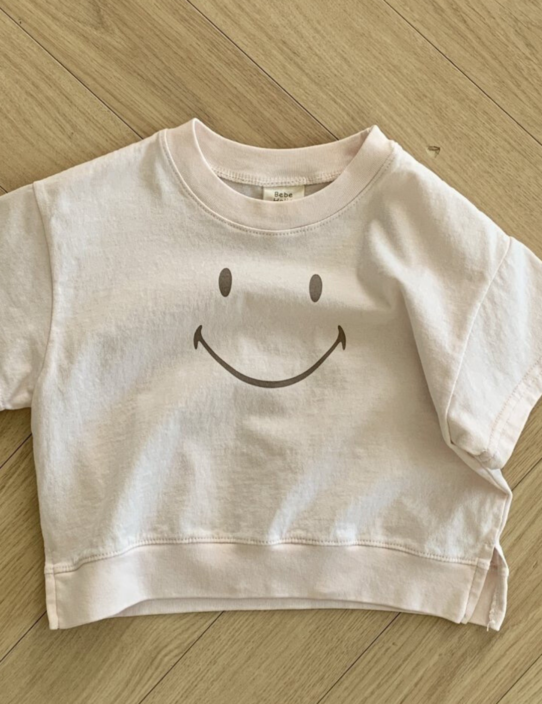 Ein blassrosa T-Shirt aus weicher Baumwolle liegt auf einem Holzboden. Das Bebe Holic Smile Tee hat in der Mitte ein einfaches schwarzes Smiley-Design. Der Boden besteht aus hellem Holz mit einem Fischgrätenmuster.