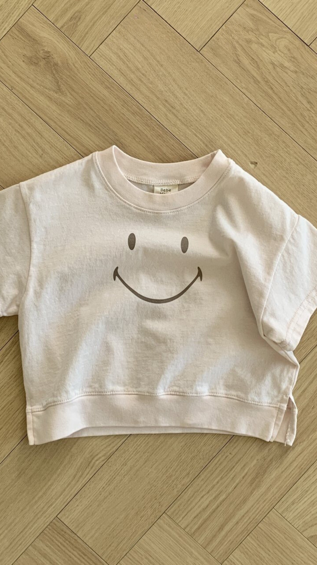 Ein blassrosa T-Shirt aus weicher Baumwolle liegt auf einem Holzboden. Das Bebe Holic Smile Tee hat in der Mitte ein einfaches schwarzes Smiley-Design. Der Boden besteht aus hellem Holz mit einem Fischgrätenmuster.