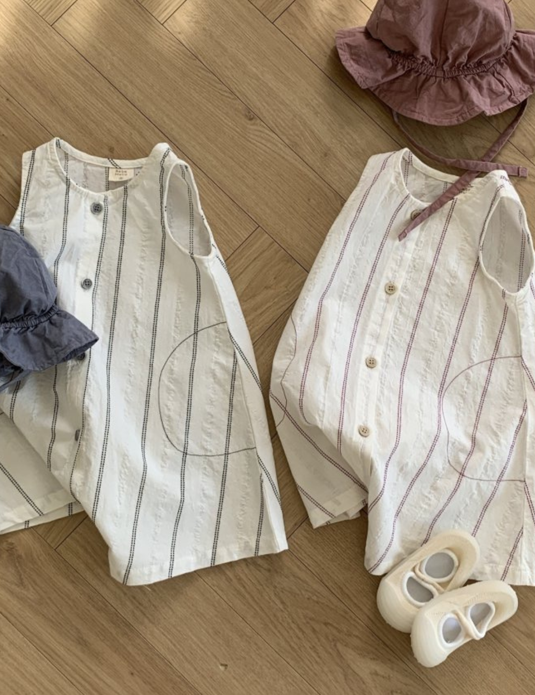 Zwei Sommeroutfits auf einem Holzboden, bestehend aus gestreiften ärmellosen Oberteilen mit passenden Hüten und weißen Sandalen daneben. Die Shirts haben ein Streifendesign mit leichten rosa Akzenten.
Produktname: Daisy ST Bodysuit
Markenname: Bebe Holic