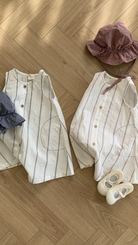 Zwei Sommeroutfits auf einem Holzboden, bestehend aus gestreiften ärmellosen Oberteilen mit passenden Hüten und weißen Sandalen daneben. Die Shirts haben ein Streifendesign mit leichten rosa Akzenten.
Produktname: Daisy ST Bodysuit
Markenname: Bebe Holic