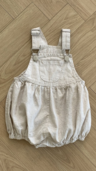 Ein hellbeiger Baby-Strampler aus Leinen mit Schnallen von Bebe Holic mit verstellbarem Träger und einer einzelnen Vordertasche liegt auf einem Holzboden. Der Strampler hat elastische Beinöffnungen, eine lockere, bauschige Passform und ist aus atmungsaktiver Baumwolle gefertigt.