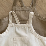 Zwei Twill-Bodysuits mit Taschen von Bebe Holic liegen auf einem hellen Fischgrätmusterboden aus Holz. Ein Bodysuit ist hellbeige mit einer kleinen Vordertasche, der andere ist dunkler beige und hat eine schlichte Vorderseite. Beide Bodysuits sind aus weicher Baumwolle gefertigt und haben verstellbare Träger für eine einfache Anpassung.