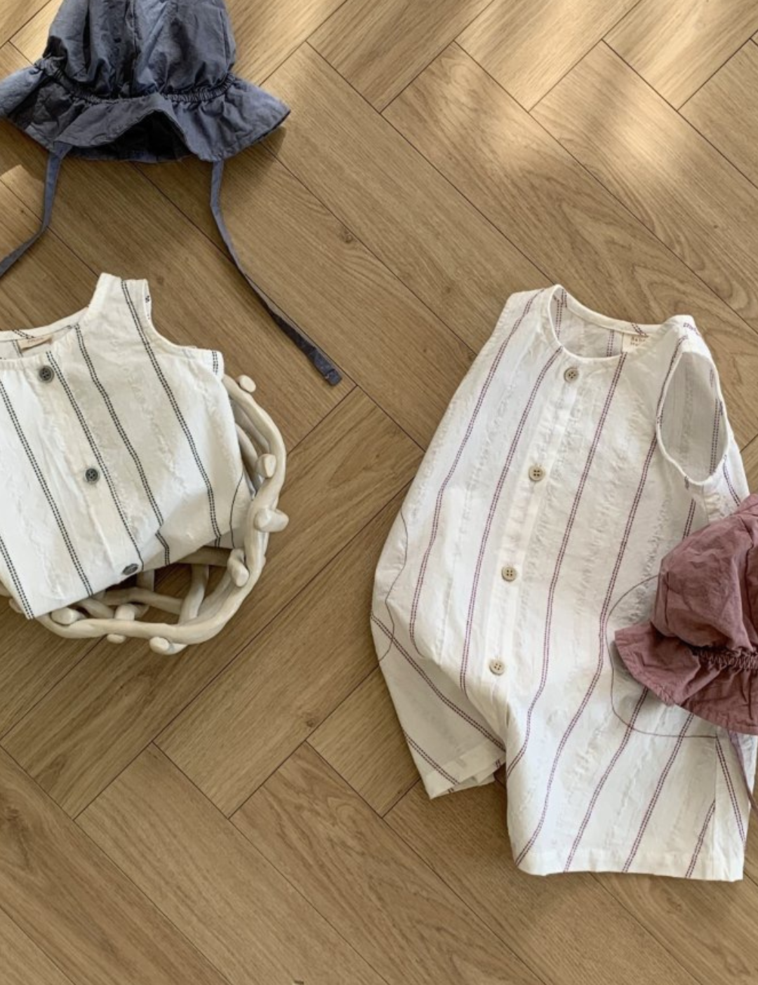 Ein gestreiftes Korsett liegt auf einem Holzboden neben einem Paar ähnlich gestalteter Bebe Holic Daisy ST Body Suits, überall sind ein Hut aus grauem Stoff und ein weinrotes Stück Stoff verstreut.