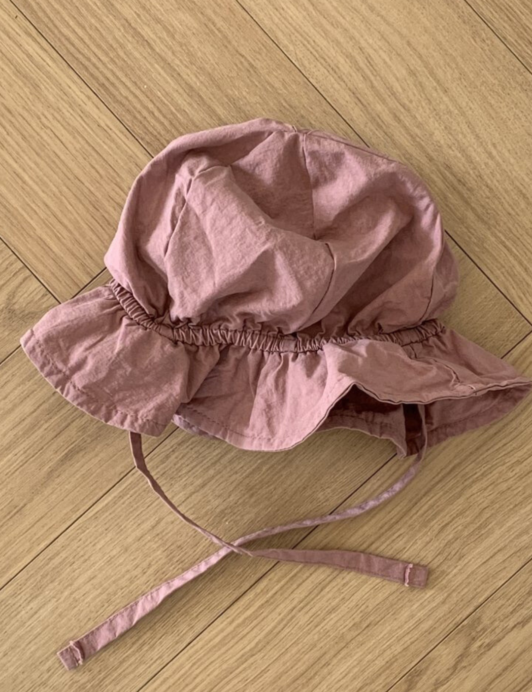 Ein Crunch Bucket Hat von Bebe Holic mit Kinnriemen liegt auf einem hellbraunen Holzboden. Der Hut, Teil der Kinderhüte-Kollektion, hat eine faltige Textur und ein Gummiband um die Kronenbasis und fügt sich nahtlos in jede natürliche Farbpalette ein.