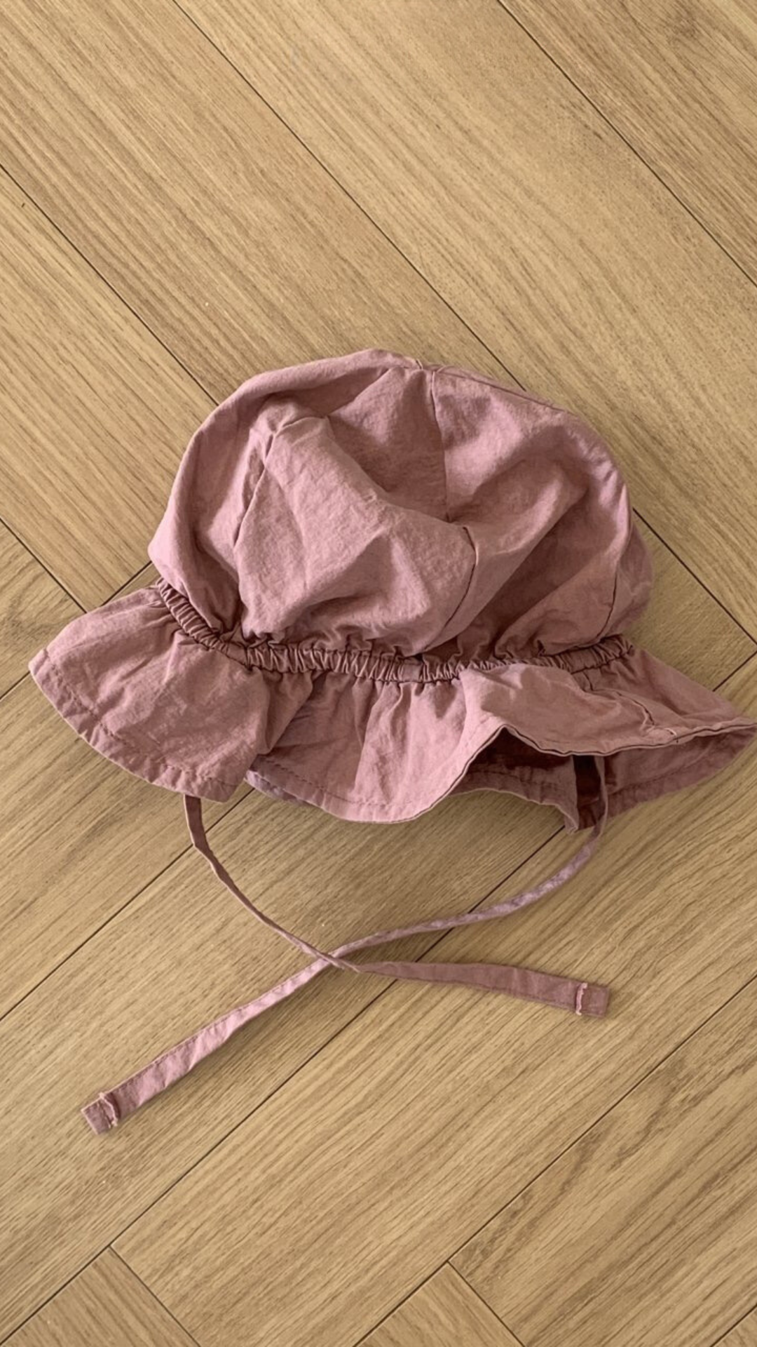 Ein Crunch Bucket Hat von Bebe Holic mit Kinnriemen liegt auf einem hellbraunen Holzboden. Der Hut, Teil der Kinderhüte-Kollektion, hat eine faltige Textur und ein Gummiband um die Kronenbasis und fügt sich nahtlos in jede natürliche Farbpalette ein.