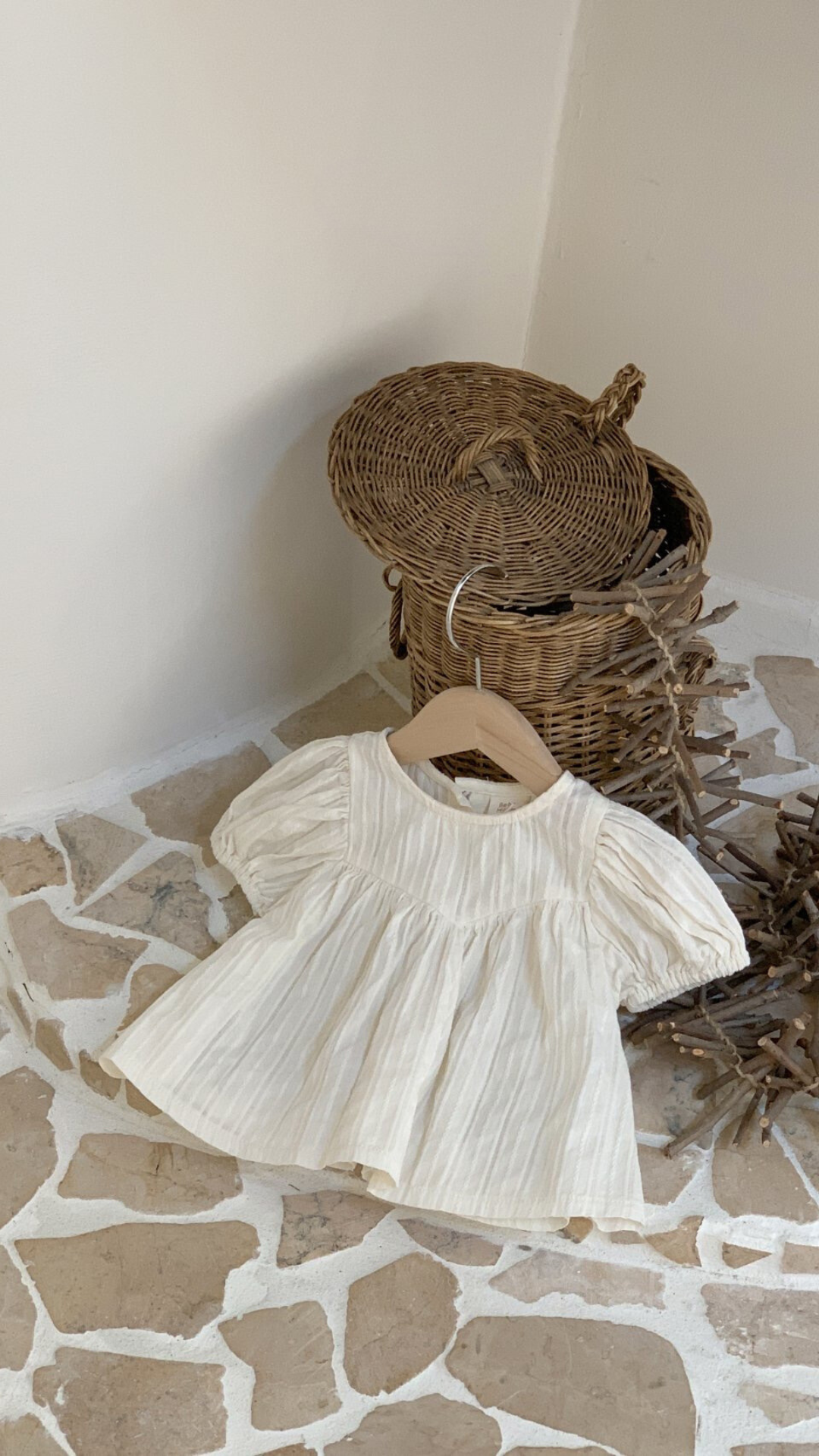 Eine kleine, schlichte weiße Raoul-Bluse von Bebe Holic mit luftiger Ärmelgestaltung liegt auf einem Steinboden neben einem Weidenkorb und einem Zweigbündel, in einem sanft beleuchteten Raum mit weißen Wänden.