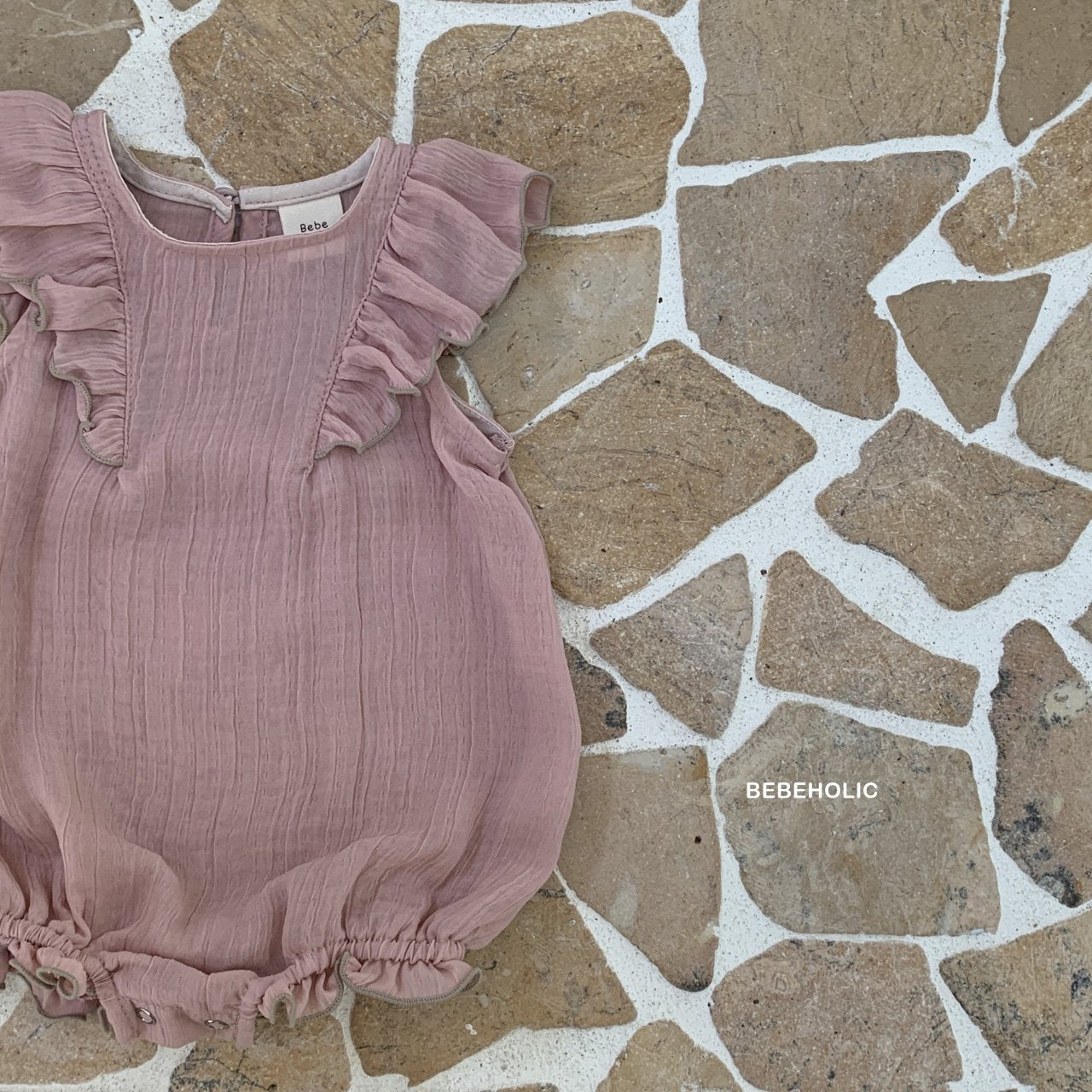 Ein rosafarbener Bebe Holic Lily Wing Bodysuit mit romantischen Rüschenärmeln, flach auf einem beigen Steinfliesenhintergrund mit weißem Fugenmuster gelegt. Die Marke „Bebe Holic“ ist sichtbar.