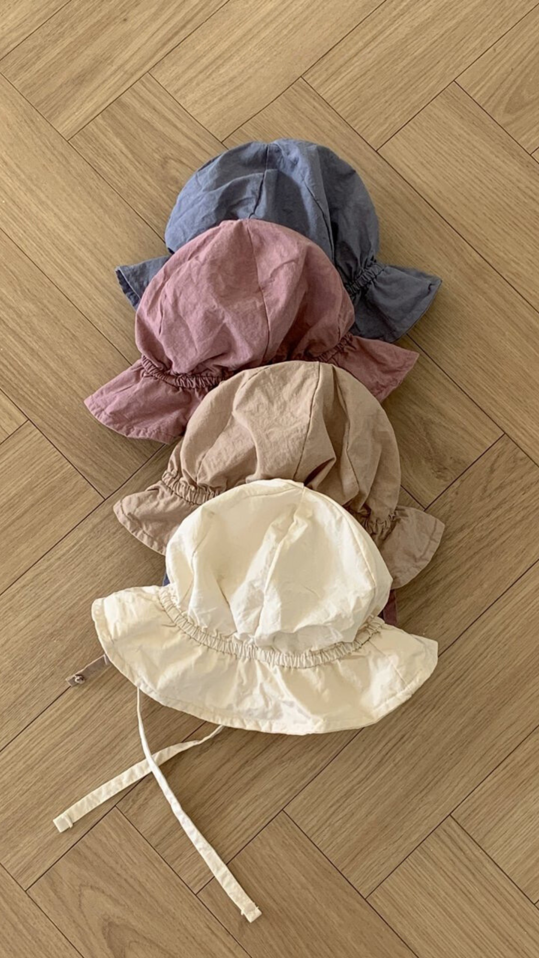 Fünf Crunch Bucket Hats in einer natürlichen Farbpalette (Blau, Rosa, Braun, Beige und Creme) sind ordentlich auf einem hölzernen Boden im Fischgrätenmuster gestapelt. Der cremefarbene Hut oben hat zwei dünne Riemen. Entdecken Sie die bezaubernde Auswahl von Bebe Holic für Ihre Kleinen.