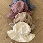 Vier Crunch Bucket Hats in verschiedenen gedeckten Tönen (Blau, Mauve, Beige und Creme) von Bebe Holic sind ordentlich auf einem hellen Holzboden mit Fischgrätenmuster gestapelt, wodurch ihre natürliche Farbpalette hervorgehoben wird.