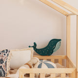 Ein gemütliches, helles Bett mit Holzrahmen in einem Kinderzimmer ist mit Kissen in verschiedenen Mustern, einem blau-weißen Ball und einer an der Wand montierten Little Lights Whale Lamp | Galaxy Teal von Little Lights dekoriert. Ein Weidenkorb ist teilweise im Vordergrund sichtbar und ergänzt die verspielte Kinderzimmerdekoration.