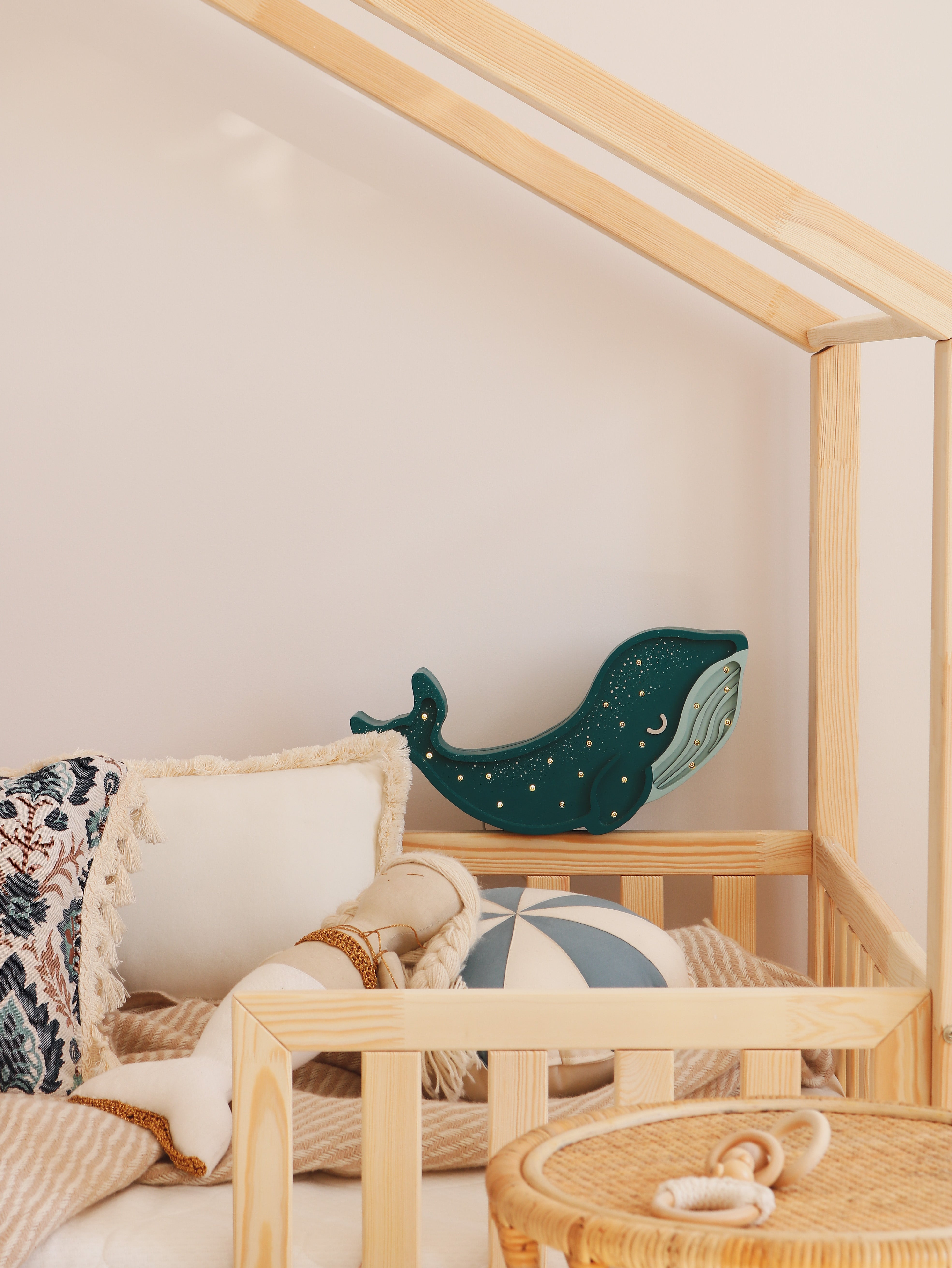 Ein gemütliches, helles Bett mit Holzrahmen in einem Kinderzimmer ist mit Kissen in verschiedenen Mustern, einem blau-weißen Ball und einer an der Wand montierten Little Lights Whale Lamp | Galaxy Teal von Little Lights dekoriert. Ein Weidenkorb ist teilweise im Vordergrund sichtbar und ergänzt die verspielte Kinderzimmerdekoration.