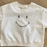 Ein weißes, kurzes Sweatshirt aus weicher Baumwolle mit einer einfachen, großen Smiley-Grafik auf der Vorderseite. Das Smile Tee von Bebe Holic liegt flach auf einem hellen Holzboden im Fischgrätenmuster.