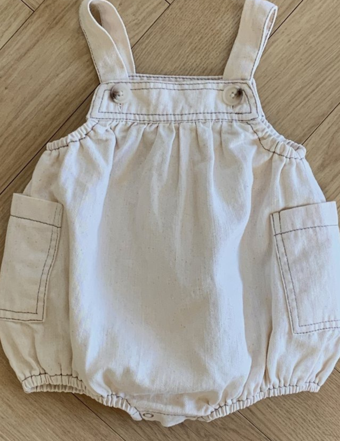 Ein hellbeiger Denim Pocket Body Suit von Bebe Holic mit verstellbaren Trägern und Taschendetails wird auf einem Holzboden ausgestellt. Der Strampler weist gerafften Stoff an der Taille und den Beinöffnungen auf, wodurch ein bauschiges Aussehen entsteht. An den verstellbaren Trägern sind zwei Knopfverschlüsse sichtbar, die Bewegungsfreiheit gewährleisten.