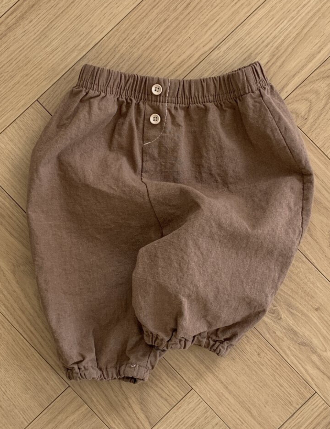 Ein Paar braune Shorts in Kleinkindergröße mit elastischer Taille und zwei Knöpfen auf der Vorderseite. Die leichten Bebe Holic Natural Pants haben elastische Bündchen an den Beinöffnungen. Diese vielseitigen Must-have-Shorts werden flach auf einem Holzboden ausgebreitet.