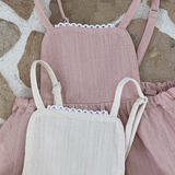 Drei pastellfarbene Bebe Holic-Kinderkleider mit Rüschenträgern und Spitzendetails, vertikal auf einem strukturierten Steinhintergrund angeordnet. Diese aus leichten Stoffen gefertigten Sommerkleider sind perfekt für warmes Wetter.