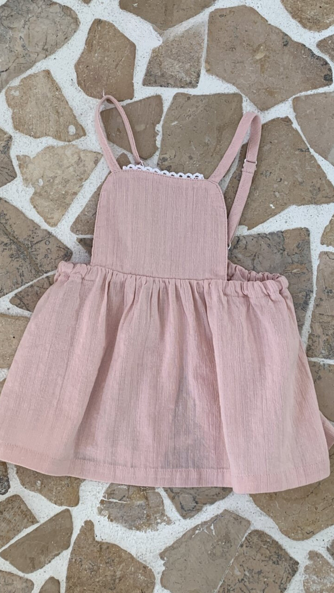 Ein rosafarbener Bebe Holic Lagenrock für Kleinkinder mit Spaghettiträgern und gerafftem Rock, präsentiert auf einem gemusterten Fliesenboden. Das Kleid hat einen zarten Spitzenbesatz über der Brust.