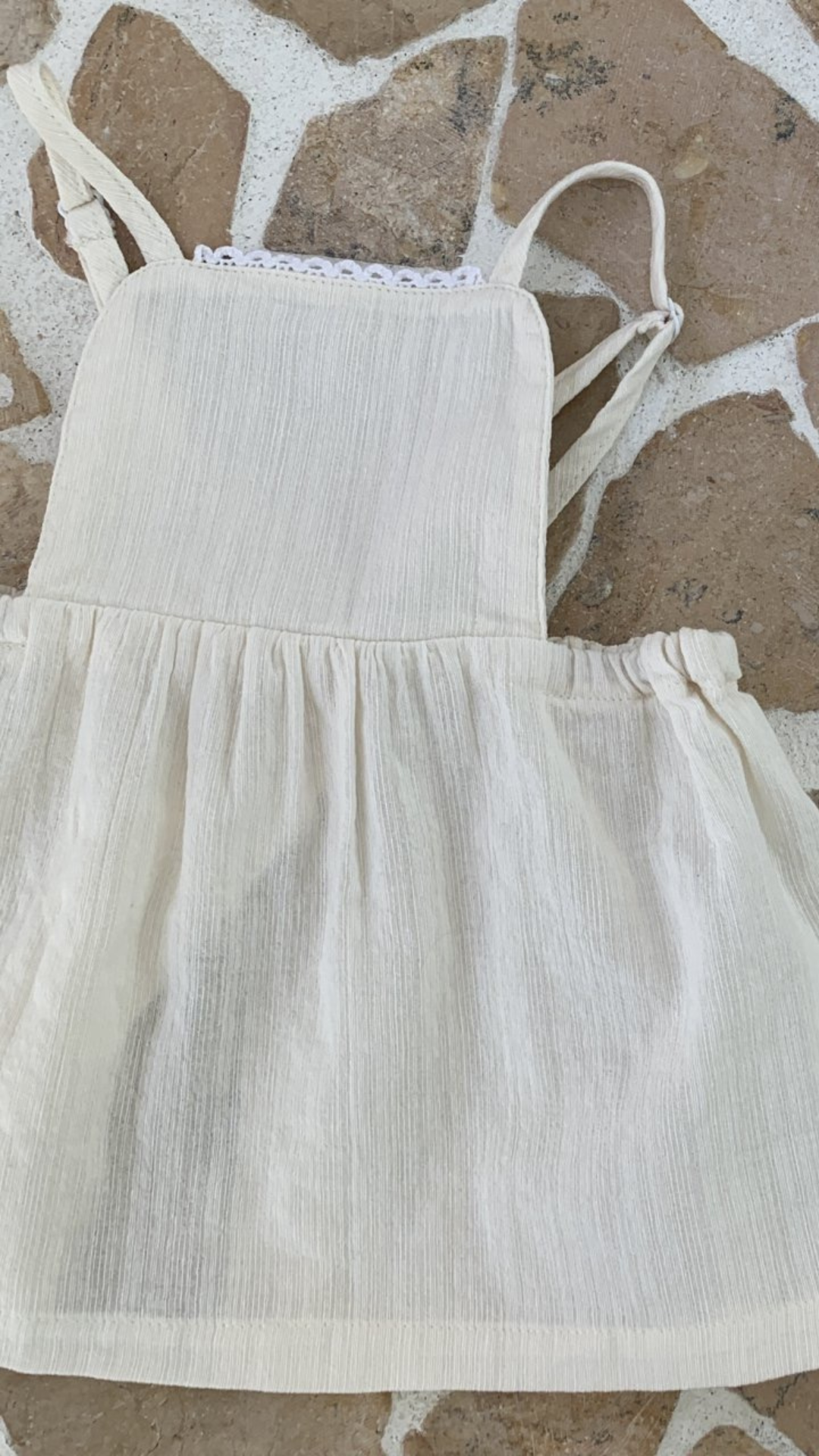 Eine weiße, strukturierte Schürze mit Latz und geraffter Taille, präsentiert vor einem Hintergrund aus Steinfliesen aus leichten Stoffen, aus der Layered Skirt-Kollektion von Bebe Holic.