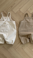 Zwei Twill-Bodysuits mit Taschen von Bebe Holic, einer weiß und einer beige, liegen flach auf einem Holzboden. Jeder hat eine Tasche auf der Vorderseite.