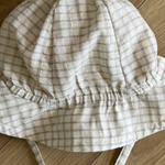 Ein beige karierter Bucket Hat mit breiter Krempe und Kinnriemen, der an traditionelle Karo-Kinderhüte erinnert, liegt flach auf einem Holzboden. Der Stoff hat ein weiches, gitterartiges Muster mit hellbraunen Linien für optimalen Sonnenschutz. Die Krempenränder sind leicht gebogen. Dies ist der Yogurt ST Bucket Hat von Bebe Holic.